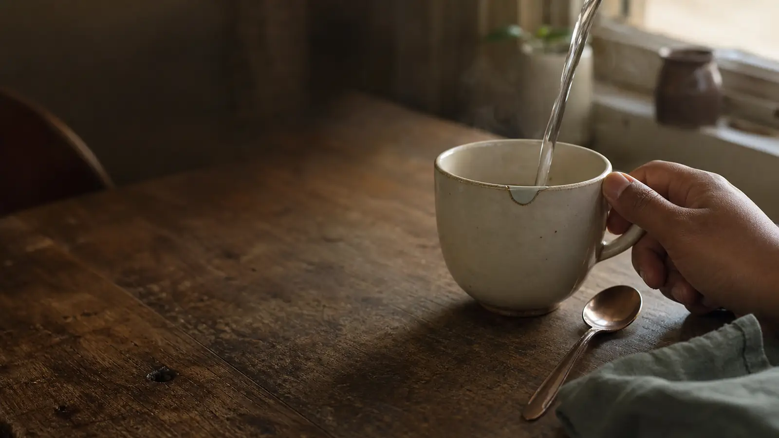 Water pours into a repaired cream cup while a brown hand steadies it beside a spoon. The ordinary object is shown through the relations that make its role visible: holding, pouring, gripping, stirring, and bearing a history.